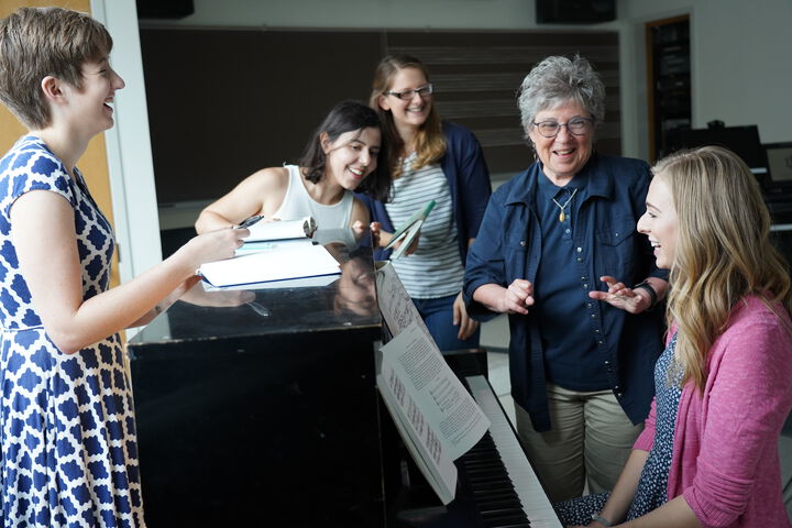 A student seated at the piano with a professor and other music students gathered around, everyone smiling.